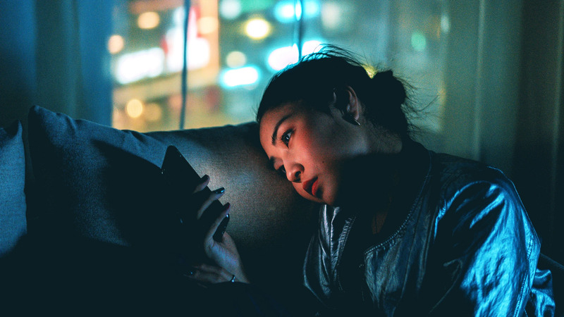 Person sitting on a couch in a dim room, holding a smartphone, with city lights visible through a window.